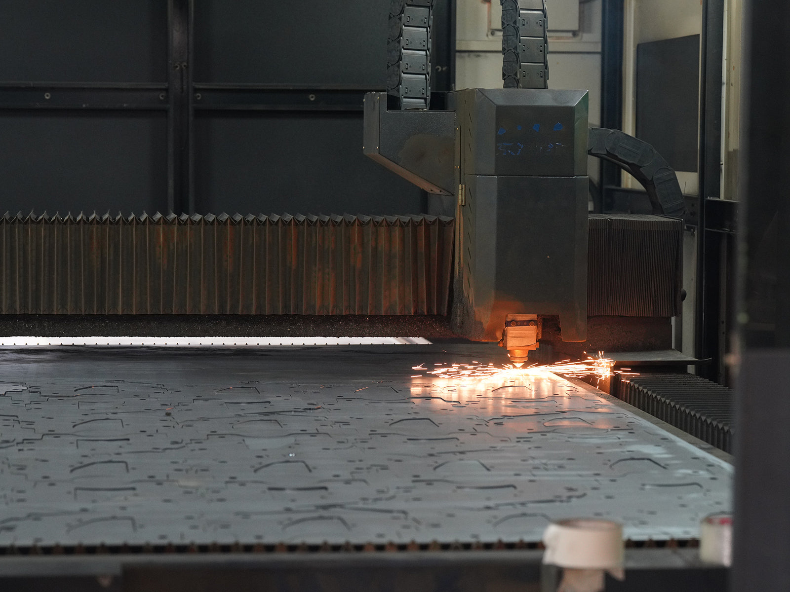 A large industrial plasma cutting machine in operation, cutting a sheet of metal with bright sparks flying at the intersection of the blade and the material.
