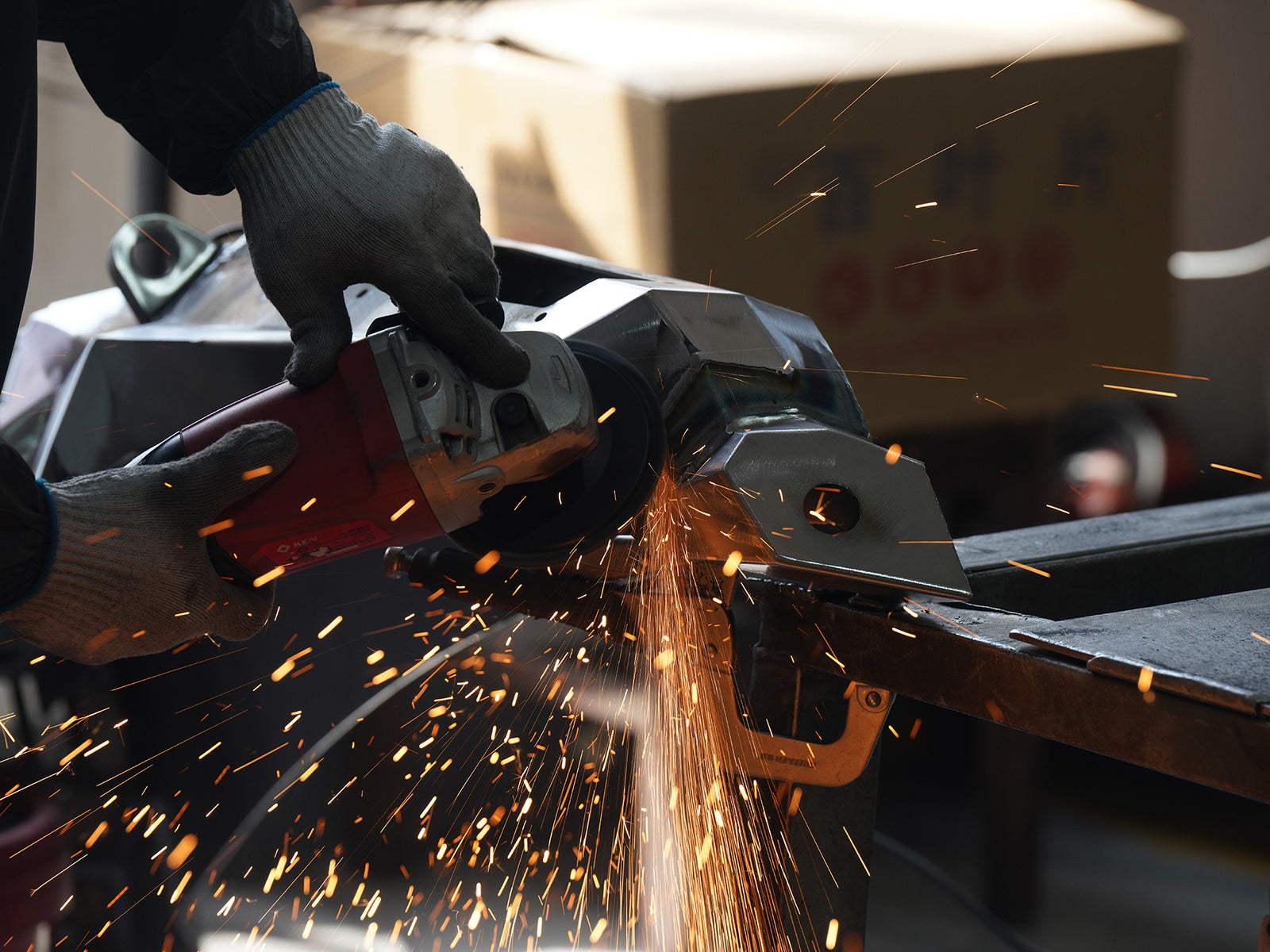 A person wearing gray work gloves operates a red and silver angle grinder, creating a shower of bright orange sparks while cutting a metal frame in an industrial setting.