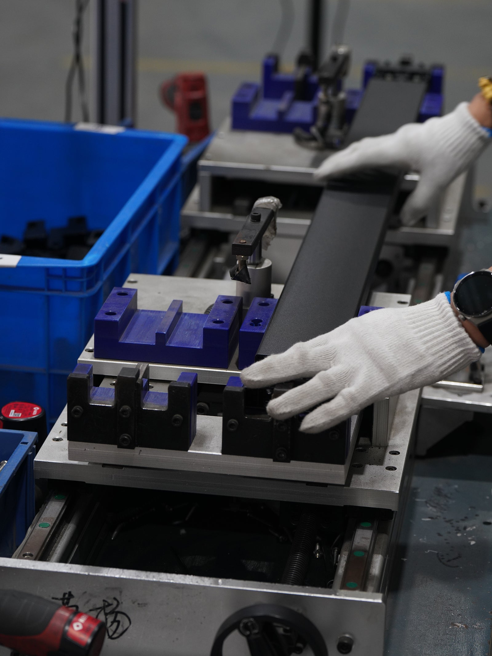 A close-up of a person wearing white knit work gloves handling a long black rectangular component on an industrial metal machine, with blue plastic bins and other machinery visible in the workshop.